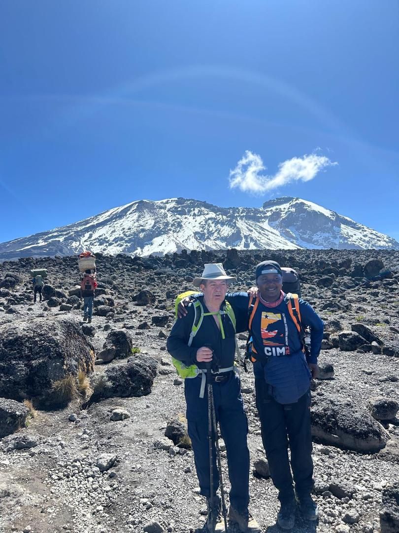 Joseph, on the right in a backwards cap, with his arm around an older hiker on a rocky plain below a snow-covered summit, porters carrying loads behind