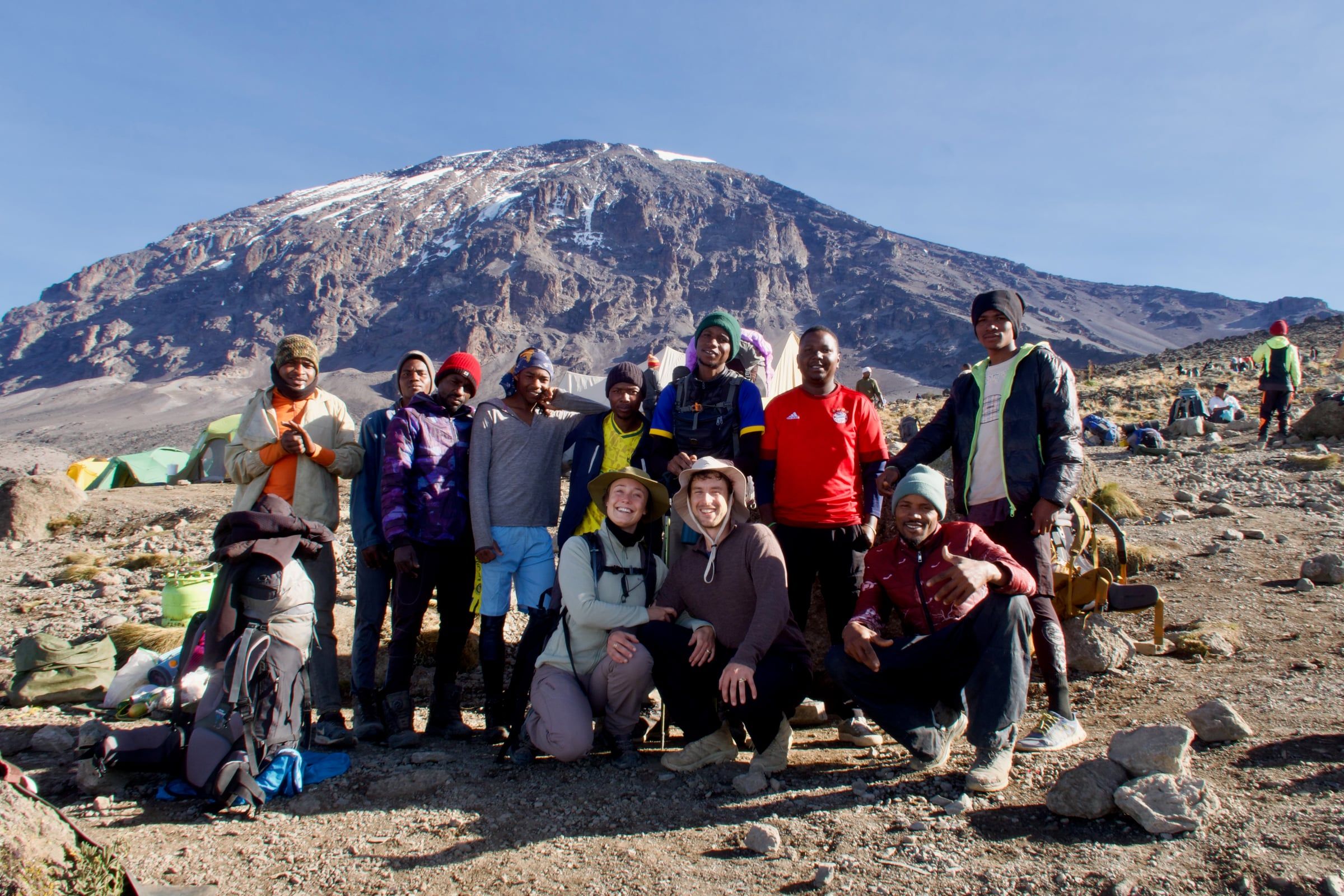 Two smiling climbers crouch with their mountain crew on rocky ground below Kilimanjaro's snow-streaked summit cone. Joseph stands at the back in a red football shirt
