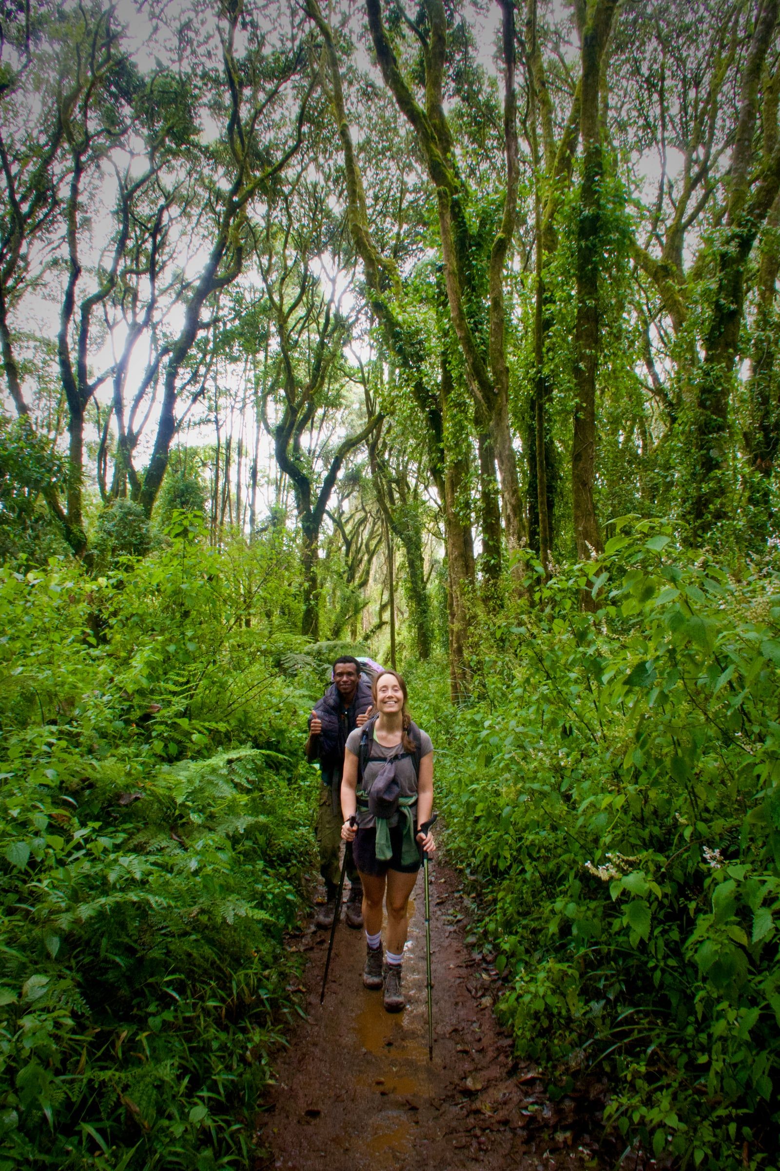 A smiling hiker with trekking poles on a muddy rainforest trail, a crew member behind her giving two thumbs up