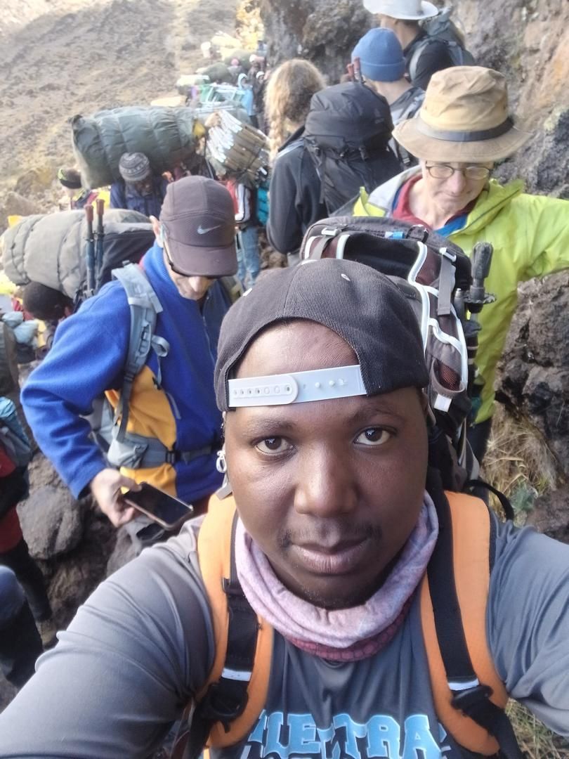 Selfie of Joseph in a backwards cap, with hikers and porters carrying loads queueing on a narrow rocky trail behind him