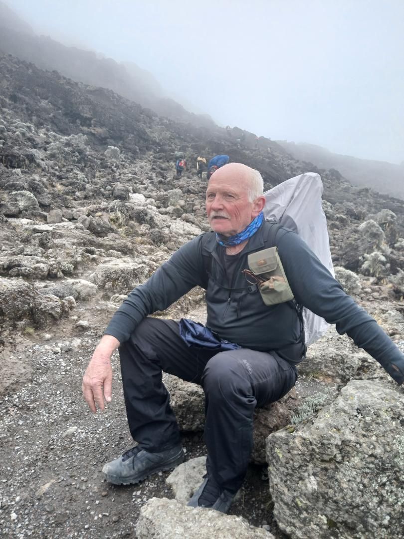 An older hiker rests on a rock on a misty, stony slope while porters carry loads up the trail behind him