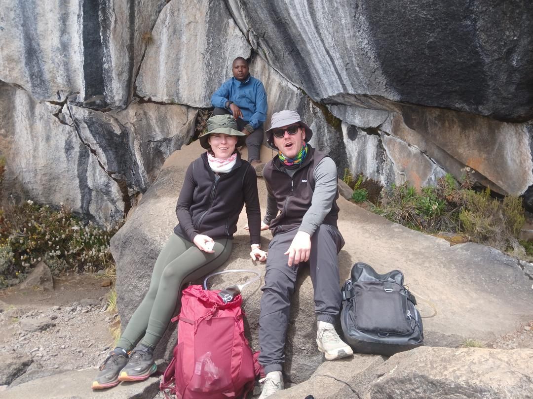 Two hikers rest with their daypacks under a large overhanging boulder while a guide sits on the rock behind them