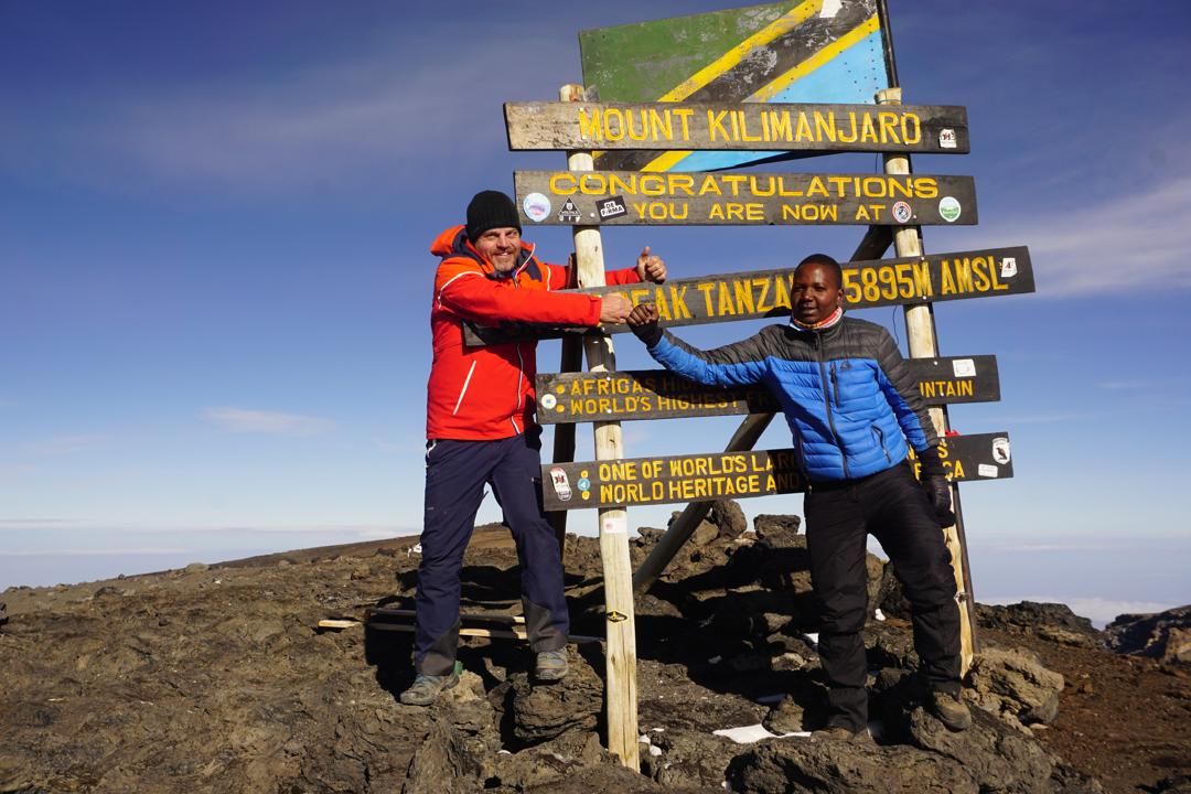 Joseph, on the right in a blue jacket, bumps fists with a climber at the Mount Kilimanjaro summit sign reading 5895M AMSL, under a clear blue sky above the clouds
